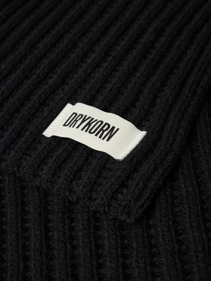 Produktfoto, schwarzer Grobstrick Schal mit weißem Label-Detail, Modell 'NUMAS' von DRYKORN, Material-Mix mit Alpaka-Anteil, Detailaufnahme