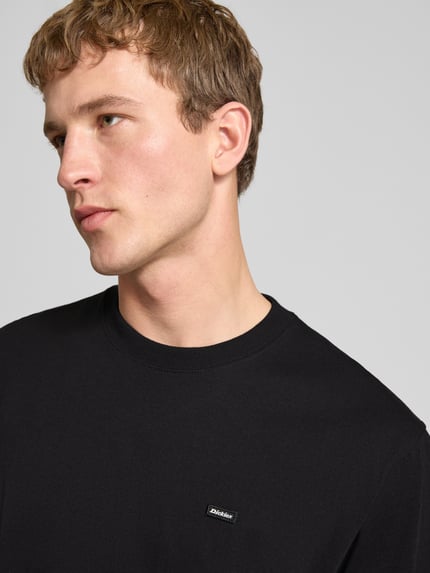 Męski model nosi T-Shirt mit Label-Detail und Rundhalsausschnitt od Dickies, bawełna, krój relaxed fit, czarny kolor, zbliżenie na górę ciała. Długość pleców w rozmiarze M: 70 cm.