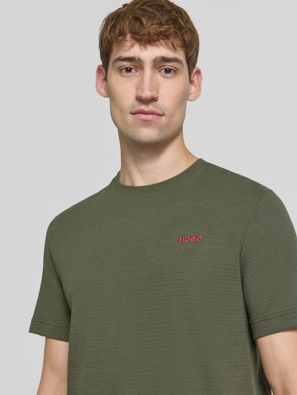 Männermodel trägt Regular Fit T-Shirt aus Baumwoll-Mix von HUGO in Olivgrün mit feiner Textur und rotem Logo, Nahaufnahme Oberkörper.