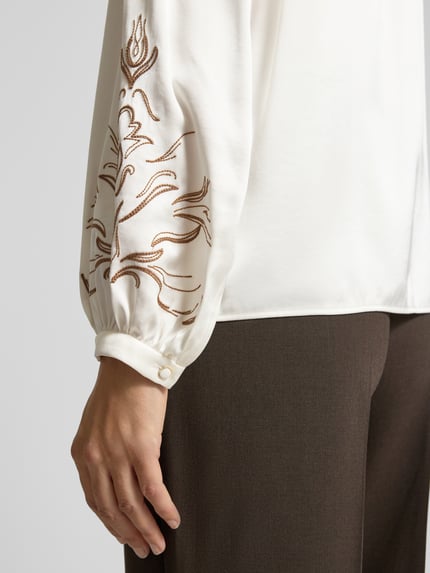 Kobieta nosi Bluzę aus Viskose-Mix mit Schlüsselloch-Ausschnitt und Stitchings von Marc Cain, kremowa i brązowa, detal rękawa, zbliżenie. Regular Fit, stójka, długie rękawy 60 cm, długość pleców 62 cm.