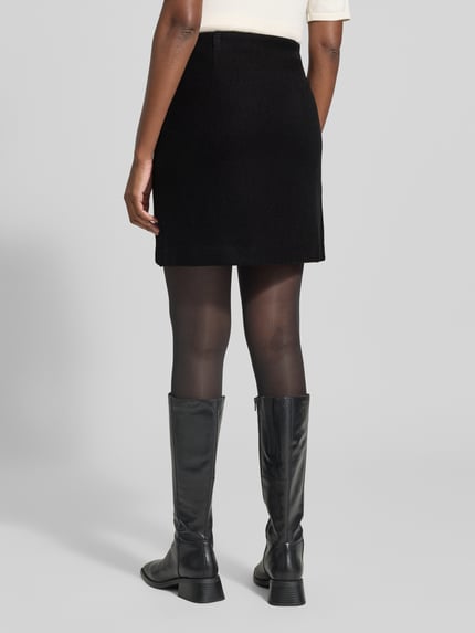 Frauenmodel trägt Ausgestellter Mini-Rock mit Nahtreißverschluss Modell 'FORTUNEALLISON' von VERO MODA in Schwarz, glatte und matte Textur, Ganzkörperansicht Rückansicht, Damen Mini-Rock von VERO MODA.