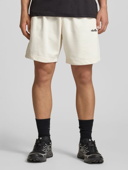 Męski model nosi Shorts mit Logo-Stitching Modell 'ZANICA' od Ellesse, bawełniany miks, krój relaxed fit, jasny kolor, elastyczny pas, kieszenie boczne, kieszeń z tyłu, logo z przodu, Szerokość w rozmiarze M: 43 cm, długość wewnętrzna nogawki M: 18 cm, zdjęcie całej sylwetki z przodu.