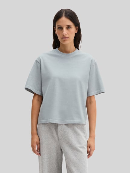 Frauenmodel trägt Boxy Fit T-Shirt aus reiner Baumwolle von Marc O'Polo, Optik: Unifarben, Passform: Boxy-Fit, Label-Schriftzug, Rundhals, Ärmelform: Kurzarm, Material: Reine Baumwolle, helle Grau- und Blautöne, glatte Textur, Ganzkörperansicht von vorne.