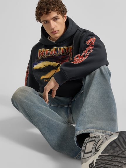 Männermodel trägt Hoodie mit Kapuze von RHUDE in Schwarz mit Label-Print und Känguru-Tasche, Reine Baumwolle, Lockerer-Schnitt, Nahaufnahme Oberkörper.
