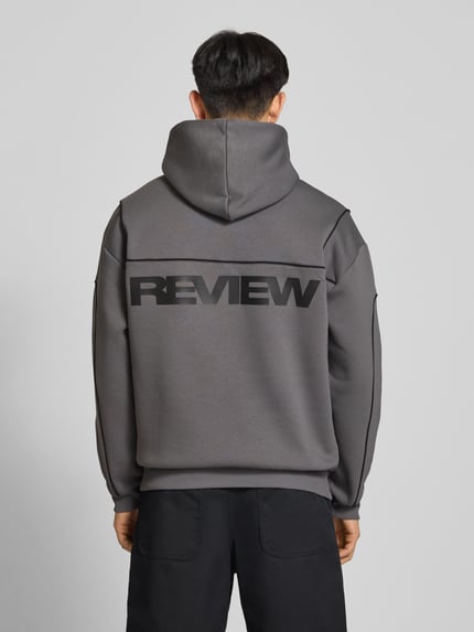 Männermodel trägt hoodie met logo en kangoeroezak van REVIEW in grijs katoenmix, regular fit, ruglengte maat M 71 cm, mouwlengte 67 cm, Ganzkörperansicht Rückansicht.