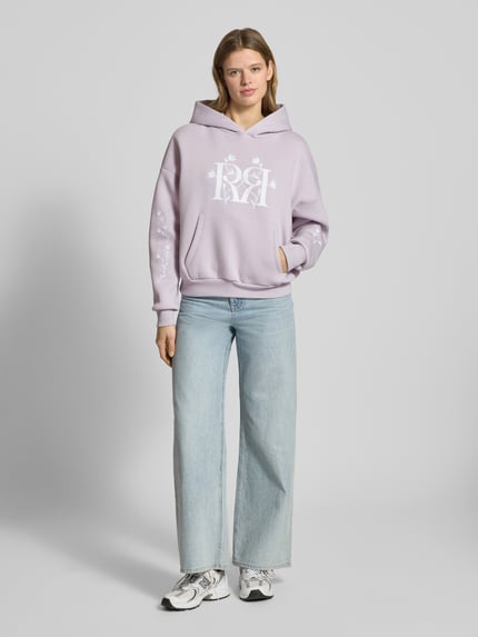 Frauenmodel trägt Relaxed Fit Hoodie mit Logo-Print von Review in Flieder, Känguru-Tasche, florales Stitching, elastische Abschlüsse, Ganzkörperansicht von vorne.