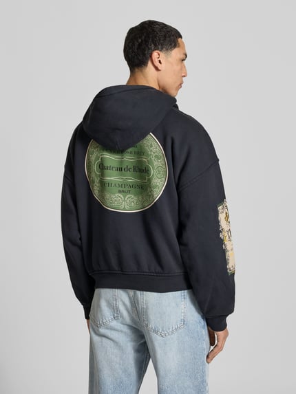 Mannenmodel draagt Oversized Hoodie aus reiner Baumwolle van RHUDE, zwart met labelprint, kangoeroezak, ribboorden, ruglengte maat M 63 cm, Ganzkörperansicht Rückansicht