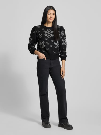 Frauenmodel trägt Regular Fit Weihnachtspullover mit Allover-Muster Modell 'NEWSNOWFROST' von Vero Moda in Schwarz mit weißen Schneeflocken, Motiv-Print, Rundhals, Langarm, gerippter Rumpfabschluss, Strick; Ganzkörperansicht von vorne.