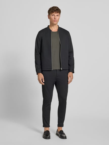 Męskimodel nosi Regular Fit Sweatjacke mit Stehkragen od Lindbergh, elastyczna mieszanka materiałów, zamek błyskawiczny, wpuszczane kieszenie, kolor czarny, długość tyłu w rozmiarze M: 69 cm, długość rękawów w rozmiarze M: 66 cm, krój regular fit, Ganzkörperansicht von vorne.