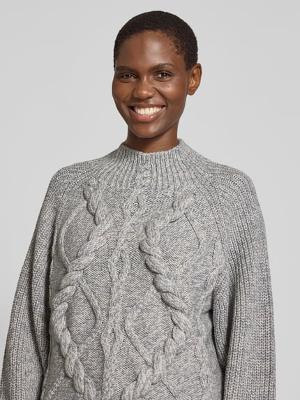 Frauenmodel trägt Pullover mit markantem Zopfmuster Modell 'Frescienne' von BOSS, Melangeoptik, hochgeschlossen, Strickstruktur, Nahaufnahme Oberkörper.