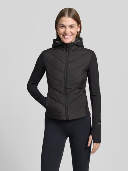 Modelka prezentuje czarną Steppjacke mit Kapuze Modell 'GRAZIA' od GUESS Activewear, krój slim fit, zamek błyskawiczny, długa rękaw, długość tyłu w rozmiarze S: 55 cm, szerokość ramion w rozmiarze S: 38 cm, szerokość w biuście w rozmiarze S: 40 cm, długość rękawa w rozmiarze S: 68 cm, widok całej sylwetki od przodu.