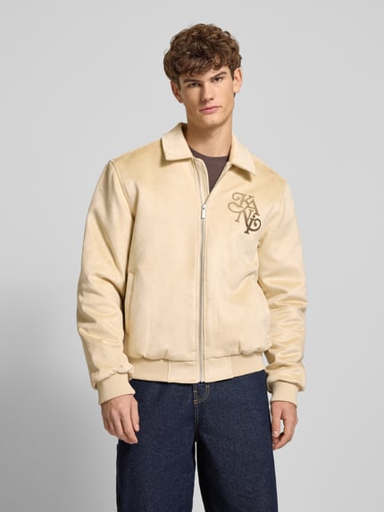 Männermodel trägt Jacke in Leder-Optik mit Label-Stitching von KARL KANI in Beige, Material-Mix mit Stretch-Anteil, Regular Fit, Reißverschluss, Umlegekragen, Ganzkörperansicht von vorne.