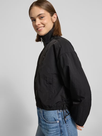 Frauenmodel trägt Relaxed Fit Blouson mit Taillenbändern von Tommy Jeans, Optik: Unifarben, Stehkragen, Reißverschluss, Logo-Stitching, Langarm, elastischer Ärmelabschluss, seitliche Eingrifftaschen mit Druckknopf, Ganzkörperansicht Rückansicht.