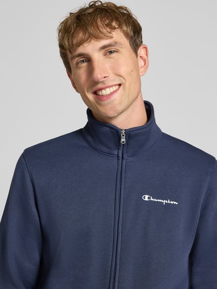 Męskimodel nosi 'Sweatanzug mit Label-Details' od Champion, kolor granatowy, mieszanka bawełny, stójka, detal z logo, zamek błyskawiczny, prosty krój, wpuszczane kieszenie, elastyczny pas, Nahaufnahme górnej części ciała.