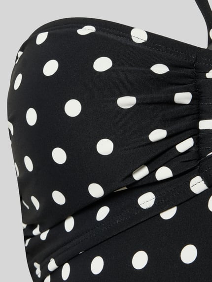 Frauenmodel trägt Bandeau-Badeanzug im Allover-Look Modell 'CLASSIC DOT BANDEAU' von Michael Kors, schwarzer Material-Mix mit Stretch-Anteil und weißen Punkten, körpernahe Passform, Nahaufnahme Oberkörper.