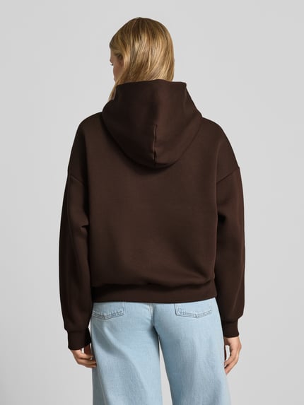 Frauenmodel trägt Relaxed Fit Hoodie aus Baumwoll-Mix mit Label-Detail von REVIEW, dunkelbraun, weiche Textur, lockere Form, Ganzkörperansicht Rückansicht