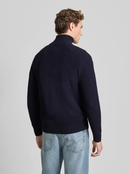Männermodel trägt Regular Fit Strickjacke aus reiner Wolle von Forét in Dunkelblau mit geripptem Stehkragen, Rückenansicht Ganzkörperansicht.
