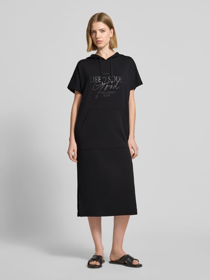 Frauenmodel trägt Sweatkleid mit Kapuze von Soyaconcept aus schwarzem Material-Mix mit Modal-Anteil, Label-Print, Gerader-Schnitt, Midikleid, Ganzkörperansicht von vorne.