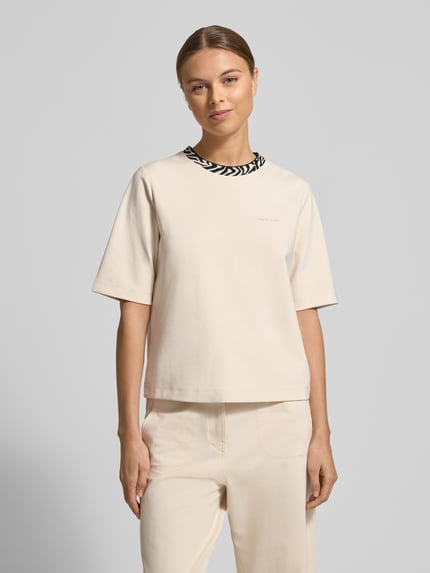 Kobiece modelka nosi damski t-shirt z mieszanki bawełny i modalu marki Marc Cain, krój regular fit, logo, okrągły dekolt, jasny kolor, gładka tekstura, całkowity widok z przodu.