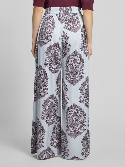 Frauenmodel trägt Wide Leg Pants mit Eingrifftaschen Modell 'Lunet' von Neo Noir, florales Muster, elastischer Bund, Ganzkörperansicht Rückansicht, Damen Wide Leg Pants, Wide Fit, französische Eingrifftaschen, Bundweite 37 cm, Innenbeinlänge 78 cm.