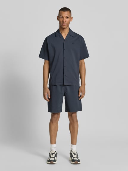 Männermodel trägt Relaxed Fit Freizeithemd aus reiner Baumwolle von Lacoste in Denim-Optik mit Crinkle-Optik, Label-Patch, Ganzkörperansicht von vorne.