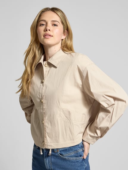 Modelka kobieca nosi Cropped Jacke mit Eingrifftaschen od Jake*s Collection, beżowa bawełna, wykładany kołnierz, listwa guzikowa, wpuszczane kieszenie, długość tyłu w rozmiarze 36: 53 cm, szerokość ramion w rozmiarze 36: 39 cm, szerokość klatki piersiowej w rozmiarze 36: 44 cm, długość rękawów w rozmiarze 36: 55 cm, cała sylwetka z przodu.