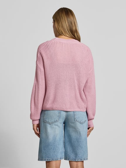 Frauenmodel trägt Boxy Fit Strickjacke mit V-Ausschnitt Modell 'CAROL' von ONLY, rosa Grobstrick, kombiniert mit hellblauer Jeansbermuda, Damen Strickjacke von ONLY, Ganzkörperansicht Rückansicht.