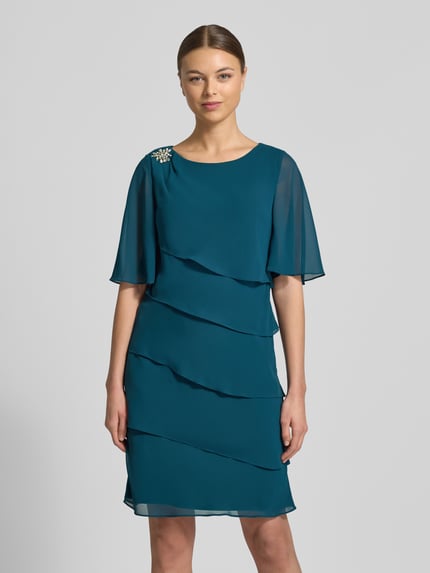 Modelka kobieca ma na sobie Relaxed Fit Kleid mit Ziersteinbesatz od Christian Berg Cocktail, turkusowy, półprzezroczysty, półrękaw, rzędy falban, okrągły dekolt, kryta halka, długość tyłu 95 cm w rozmiarze 36, szerokość w rozmiarze 36: 40 cm, długość rękawa 35 cm, widok całej sylwetki z przodu.
