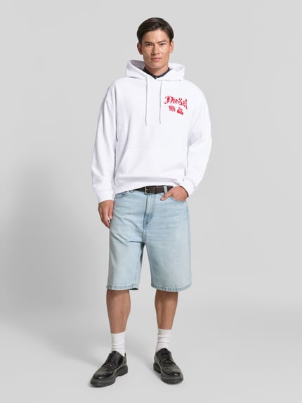 Männermodel trägt Oversized Hoodie aus reiner Baumwolle von Diesel in Weiß mit rotem Label-Print, Känguru-Tasche, Kapuze, Ganzkörperansicht von vorne.