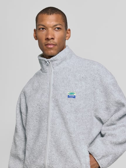 Männermodel trägt Sweatjacke Modell 'HOKTOWN' mit Logo-Patch von AMERICAN VINTAGE in Hellgrau, strukturierte Fleece-Optik, Nahaufnahme Oberkörper.