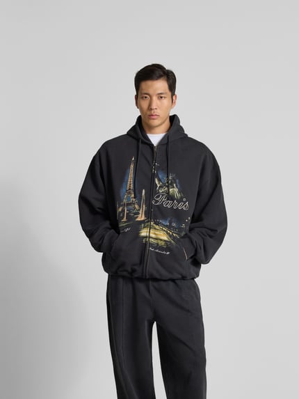 Männermodel trägt Oversized Sweatjacke mit Foto-Print von VETEMENTS aus Baumwoll-Mix, Oversized-Fit, Zweiwege-Reißverschluss, Kapuze mit Tunnelzug, dunkle Farben, weiche Textur, Rückenlänge 74 cm, Schulterbreite 68 cm, Ärmellänge 67 cm, Ganzkörperansicht von vorne.
