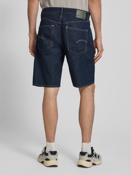 Männermodel trägt Regular Fit Jeansshorts aus reiner Baumwolle Modell 'DAKOTA SHORT' von G-Star in dunkelblauer Denim-Optik, 5-Pocket-Design, Label-Patch und Nieten, Materialverarbeitung Twill, Ganzkörperansicht Rückansicht.