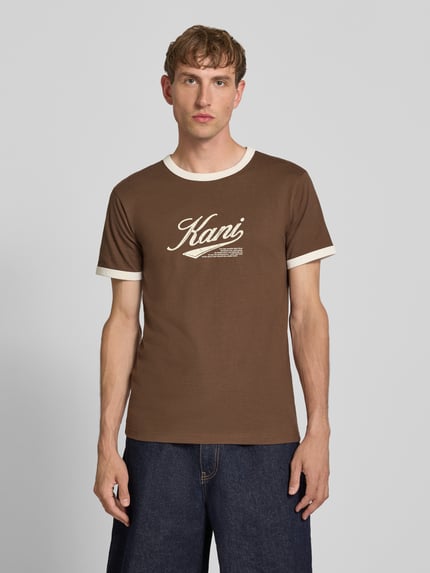 Männermodel trägt T-Shirt mit Label-Print und Rundhalsausschnitt von Karl Kani in Braun, Reine Baumwolle, Regular-Fit, Ganzkörperansicht von vorne, Rückenlänge bei Größe M 68 cm.