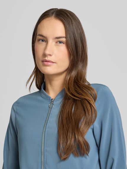 Modelka kobieca nosi Regular Fit Bomberjacke mit College-Kragen Modell 'COCO' od Vero Moda, kolor jednolity, faktura gładka, zamek błyskawiczny, długie rękawy z ściągaczami, boczne kieszenie; zbliżenie na górną część ciała.