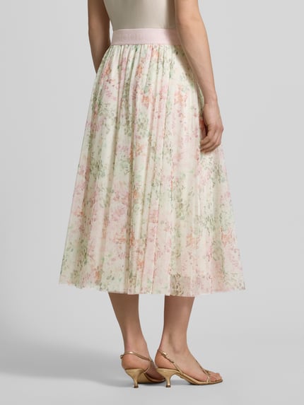 Frauenmodel trägt Midirock mit elastischem Logo-Bund von JOOP! aus Chiffon, A-Linie, Modell 'FRIDI', florales Muster, Ganzkörperansicht Rückansicht.