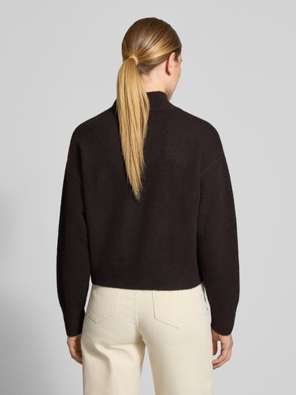 Frauenmodel trägt Turtleneck-Pullover mit Woll-Anteil und Rundhalsausschnitt von Neo Noir in Dunkelbraun, cremefarbene Hose, strukturierte Strickoptik, überschnittene Schultern, gerippte Abschlüsse, Ganzkörperansicht Rückansicht.