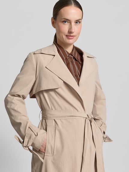 Frauenmodel trägt Trenchcoat mit Reverskragen und Bindegürtel von Zero in Beige, Taillierter Schnitt, seitliche Eingrifftaschen, Knoten-Detail am Ärmel, Nahaufnahme Oberkörper. Rückenlänge bei Größe 36: 118 cm, Schulterbreite: 39 cm, Brustweite: 48 cm, Ärmellänge: 64 cm.