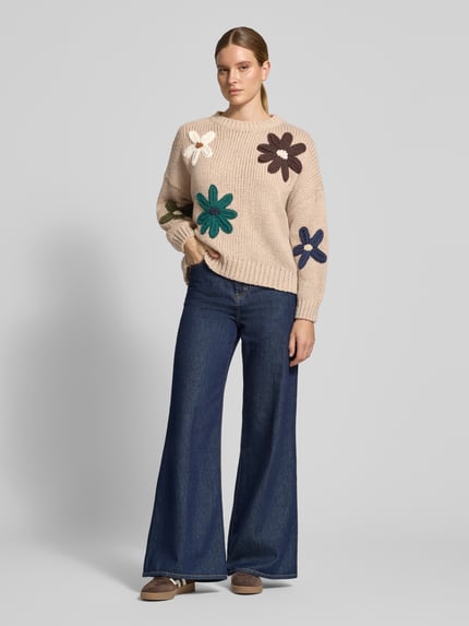 Kobiece model nosi Relaxed Fit Strickpullover mit Blumen-Stickerei od Apricot, imitacja dzianiny, krój relaxed fit, kwiatowy haft, opuszczone ramiona, krój z długim rękawem, prążkowane wykończenia, prążkowana plisa, okrągły dekolt, długość tyłu w rozmiarze S: 57 cm, długość rękawów w rozmiarze S: 44 cm, widok całej sylwetki z przodu.