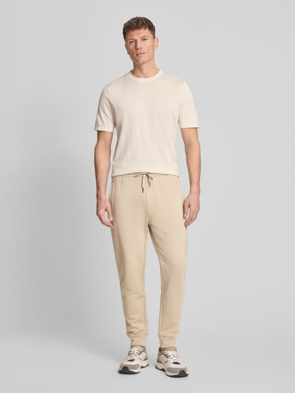 Männermodel trägt Regular Fit Sweatpants aus reiner Baumwolle von ARMANI EXCHANGE in Beige, mit elastischen Abschlüssen, Kordelzug, Seitentaschen, Gesäßtaschen, Label-Print, Innenbeinlänge 44 cm bei Größe M, Ganzkörperansicht von vorne.