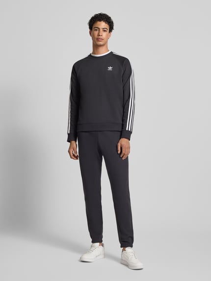 Męski model trägt spodnie dresowe z wyhaftowanym logo i elastycznym pasem od adidas Originals z bawełny, krój regular fit, długość wewnętrzna 78 cm, kolor ciemny, gładka tekstura, ganzkörperansicht von vorne.