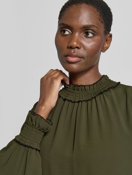 Kobieta prezentuje Loose Fit Langarmbluse mit Mock Neck od Marc Cain z szyfonu o kroju regular fit, marszczony detal na stójce, detal: marszczone mankiety, kolor oliwkowy, Długość tyłu w rozmiarze 36: 61 cm, Długość rękawów w rozmiarze 36: 63 cm, zbliżenie na górną część ciała.