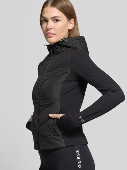 Modelka nosi czarną Steppjacke mit Kapuze Modell 'GRAZIA' von GUESS Activewear, krój slim fit, detale z matowej tkaniny, boczne kieszenie, zamek, kaptur z troczkiem, Szerokość w rozmiarze S: 40 cm, długość tyłu: 55 cm, szerokość ramion: 38 cm, długość rękawa: 68 cm, zdjęcie w półzbliżeniu z przodu.