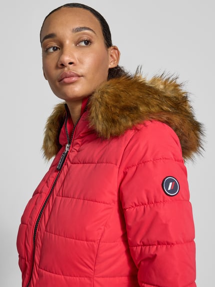 Frauenmodel trägt Steppmantel mit Zweiwege-Reißverschluss von Superdry in Rot mit Kunstfellkapuze, strukturierte Oberfläche, Nahaufnahme Oberkörper. Damen Steppmantel von Superdry, Slim Fit, Kapuze mit Kunstfell, Zweiwege-Reißverschluss, Reißverschlusstaschen.