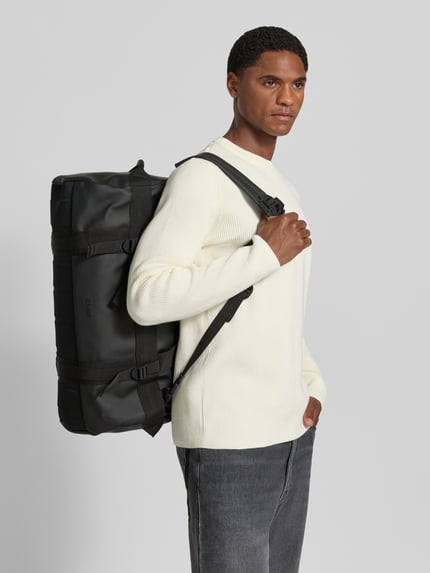 Männermodel trägt Duffel Bag mit Logo-Detail Modell 'Texel' von Rains in Schwarz, rechteckige Form, glatte Textur, Ganzkörperansicht von vorne. Hauptfach mit Reißverschluss und verstellbaren Schulterriemen, Maße 55x26x34 cm, Länge Schulterriemen 80 cm.