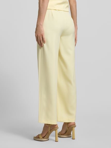 Frauenmodel trägt Relaxed Fit Hose mit gelegten Falten von Little Mistress, hellgelber Material-Mix mit Stretch, glatte Oberfläche, Ganzkörperansicht Rückansicht.