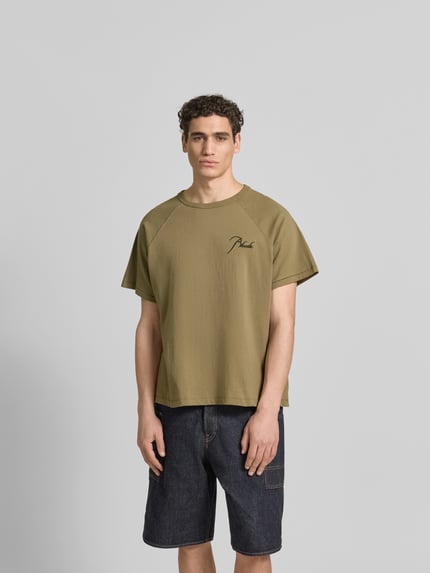 Męski model nosi Regular Fit T-Shirt aus reiner Baumwolle od RHUDE, bawełna, krój regular fit, okrągły dekolt, wyhaftowane logo, raglanowe rękawy, długość tyłu w rozmiarze M: 69 cm, oliwkowy kolor, gładka tekstura, prosty fason, widok całej sylwetki z przodu.