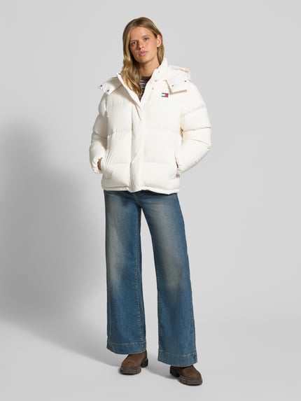 Frauenmodel trägt Regular Fit Steppjacke mit abnehmbarer Kapuze von Tommy Jeans in Weiß, kombiniert mit blauer Jeans, Ganzkörperansicht von vorne. Damen Steppjacke von Tommy Jeans.