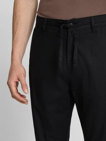 Mannelijk model draagt Relaxed Fit Hose aus Leinen-Baumwoll-Mix van s.Oliver RED LABEL, effen zwart, webstof, mid waist, knoopsluiting, zij- en achterzakken, label stitching detail, Nahaufnahme taille.