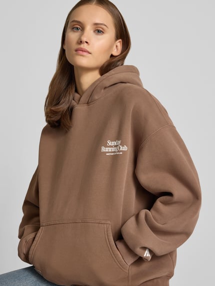 Frauenmodel trägt Relaxed Fit Hoodie aus Baumwoll-Mix von ANOTHER COTTON LAB, braun, gerippte Bündchen, Typo-Print, Nahaufnahme Oberkörper.
