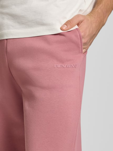 Männermodel trägt Loose Fit Sweatpants mit Seitentaschen van REVIEW in roze katoenmix met labelprint, elastische tailleband, detailopname heup.